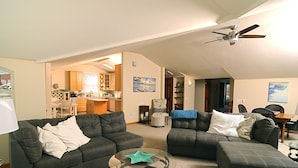 Ein geräumiger Wohnbereich mit zwei dunklen Sofas, einem Glastisch und einem Deckenventilator. Im Hintergrund ist eine Küche mit Holzausbau und einem Essbereich zu sehen.