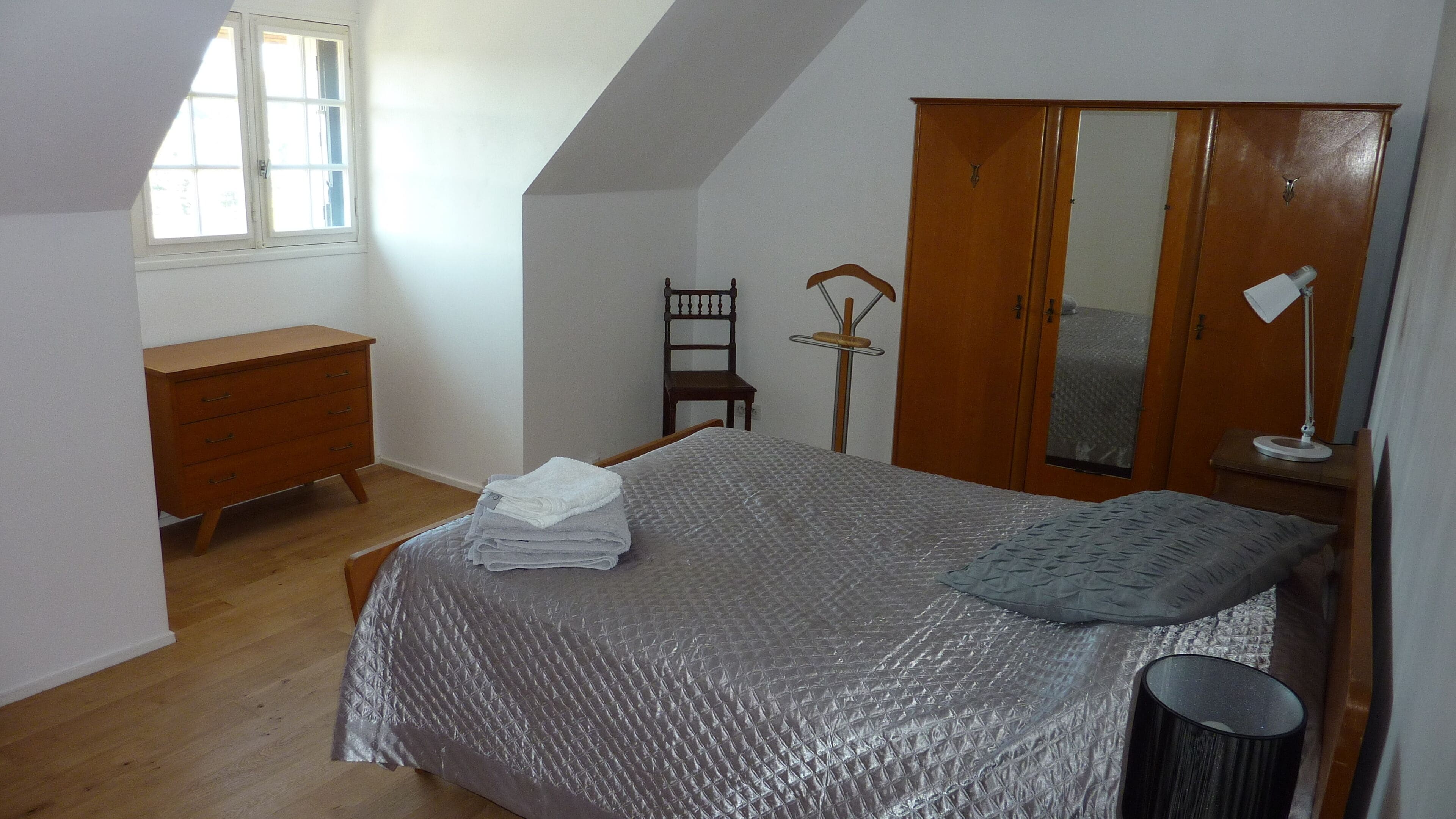 Une chambre à coucher avec un lit, une armoire en bois, une chaise, une commode et une fenêtre.