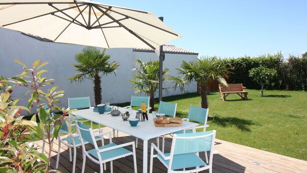 An outdoor patio with a white table and blue chairs, surrounded by greenery and a wooden deck. - House Bungalow 8p. any comfort near center and beaches, closed garden. St Denis (Saint-Denis-D'oléron)