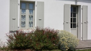 A white building with two windows and a door, surrounded by red and yellow flowering bushes. - House Bungalow 8p. any comfort near center and beaches, closed garden. St Denis (Saint-Denis-D'oléron)