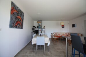 A dining area with a table and chairs, a sofa, and a bookshelf in the background. - House Bungalow 8p. any comfort near center and beaches, closed garden. St Denis (Saint-Denis-D'oléron)