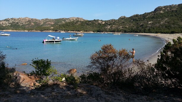 Una playa con barcos, agua cristalina y un paisaje montañoso al fondo.