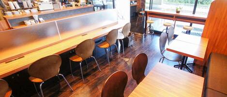 A cafe with wooden tables and chairs, a counter with a coffee machine, and large windows.