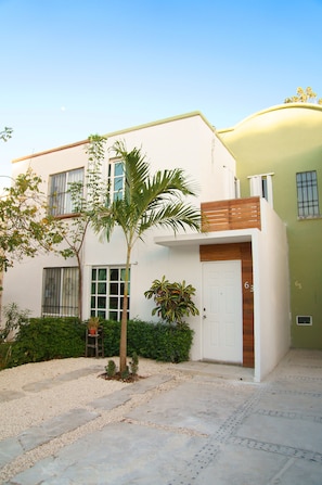 Ein modernes, zweistöckiges Haus mit weißer Fassade, einem hölzernen Balkon und einem kleinen Gartenbereich mit Palmen und Topfpflanzen.