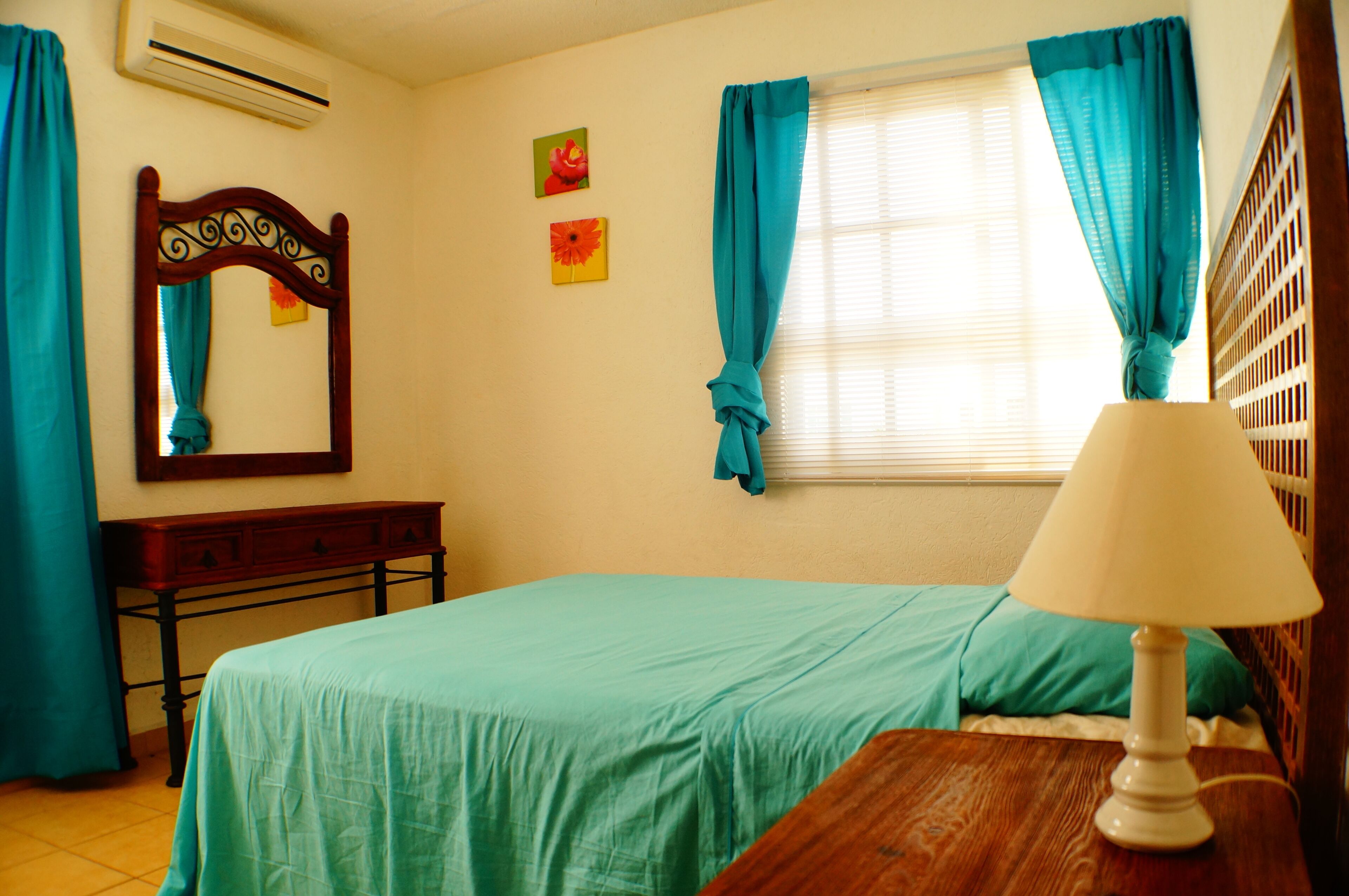 Um quarto com cama, mesa de cabeceira, abajur, cômoda, espelho e cortinas.