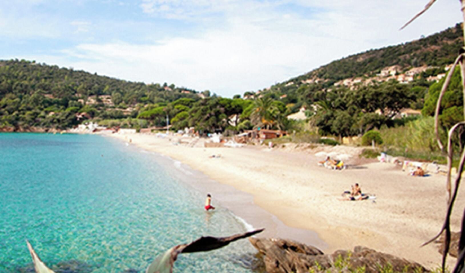 A beach with clear turquoise water, sandy shore, and a hilly backdrop with vegetation.