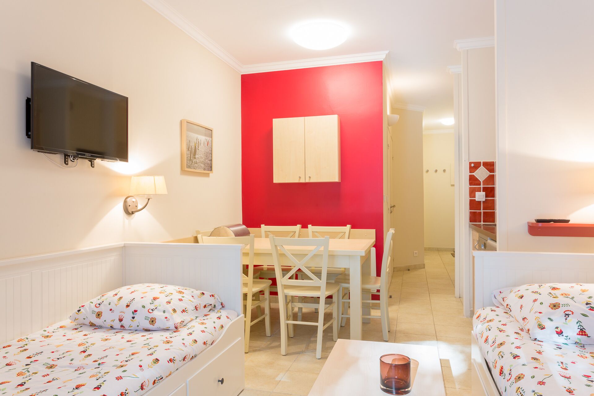 Ein Zimmer mit zwei Einzelbetten, einem Esstisch mit Stühlen, einer roten Akzentwand, einem an der Wand montierten Fernseher und einem kleinen Tisch mit einem Glas.