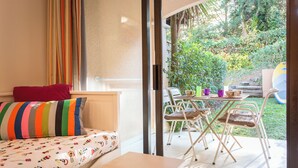 Ein Schlafzimmer mit einem Bett, einem kleinen Tisch und Blick auf einen Garten durch eine offene Tür.