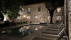 A nighttime view of a hotel courtyard with a swimming pool, lounge chairs, and a large tree. - Renovated, spacious Les toiles à Elise mansion with swimming pool (Saint-Geniès-De-Comolas)