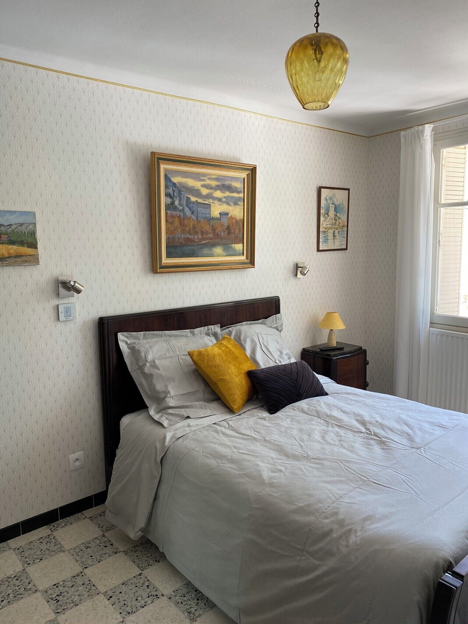 A neatly made bed with white bedding and two pillows, one yellow and one dark blue, in a room with patterned wallpaper and a painting above the bed.