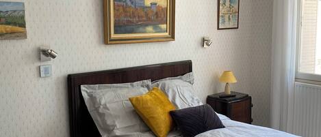A neatly made bed with white bedding and two pillows, one yellow and one dark blue, in a room with patterned wallpaper and a painting above the bed.