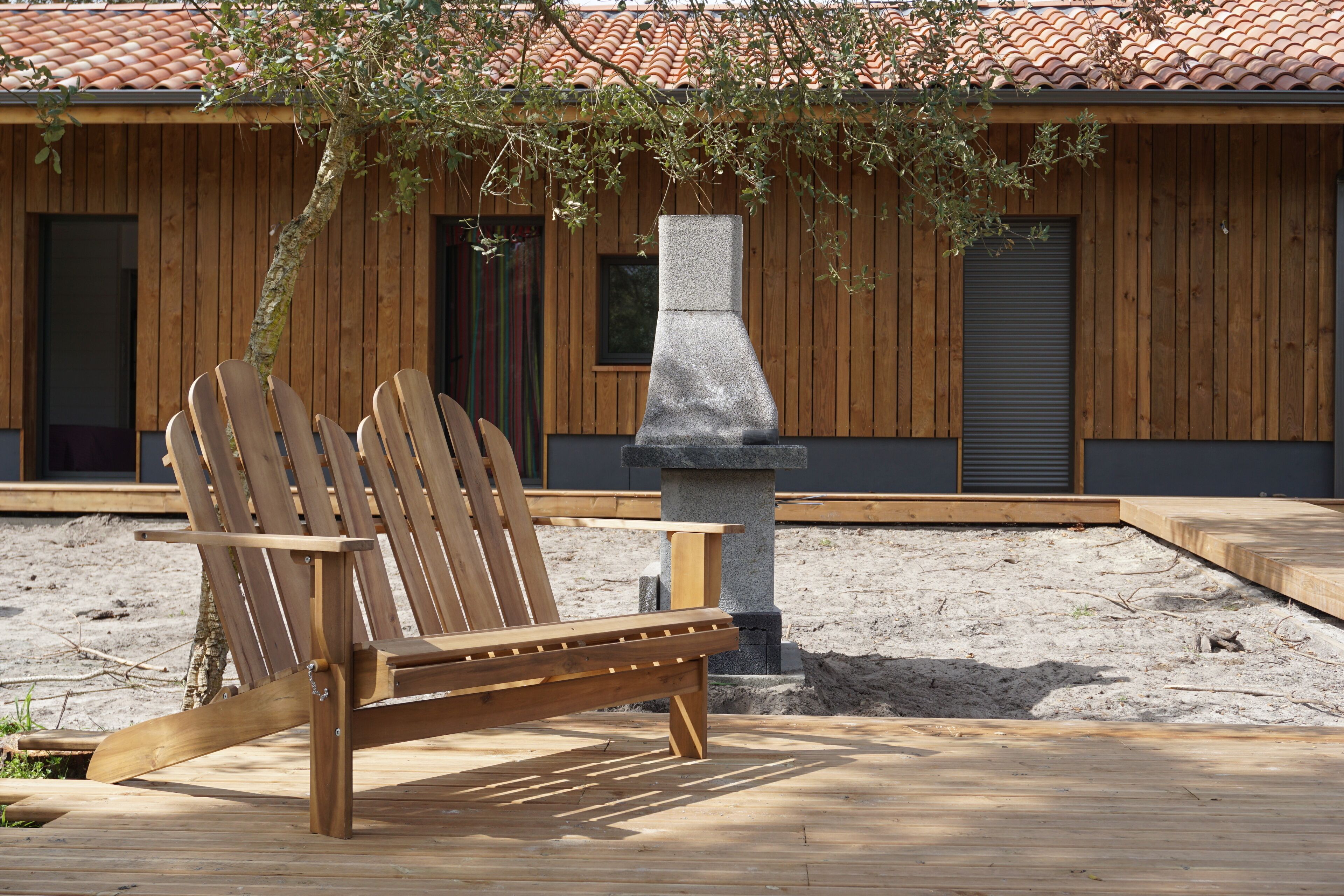 A wooden deck with a single wooden chair, a stone fireplace, and a building with wooden siding and a tiled roof.