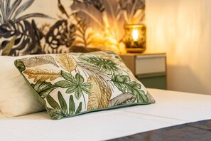 Una cama con un almohadón blanco y otro con un estampado de hojas tropicales.