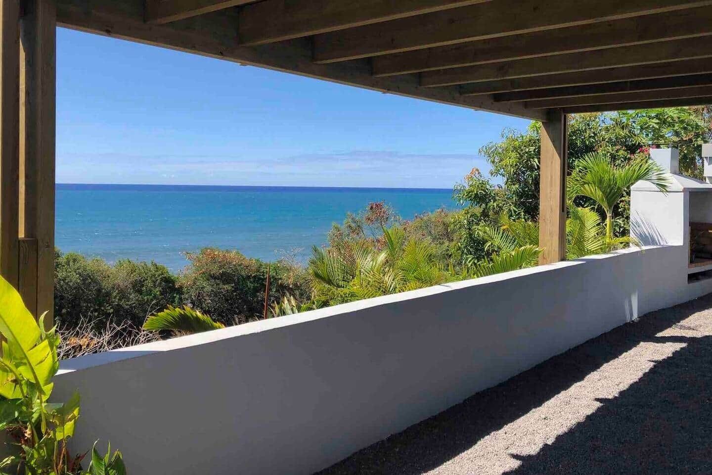Ein überdachter Außenbereich mit Blick auf das Meer, eine weiße Wand und etwas Grün.