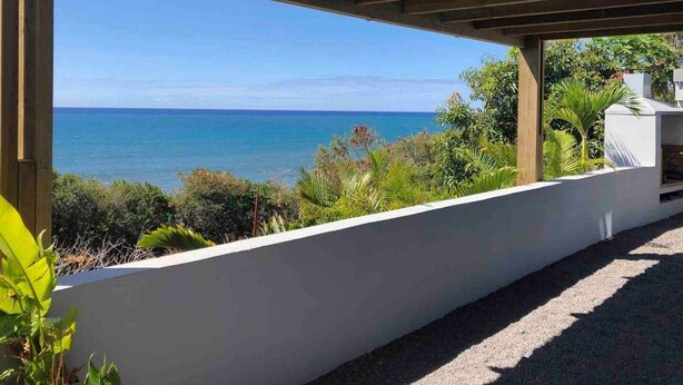 A covered outdoor area with a view of the sea, a white wall, and some greenery.
