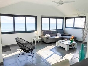 Un salón amplio con un sofá gris, una silla negra, una mesita de centro y vistas al mar a través de grandes ventanales.