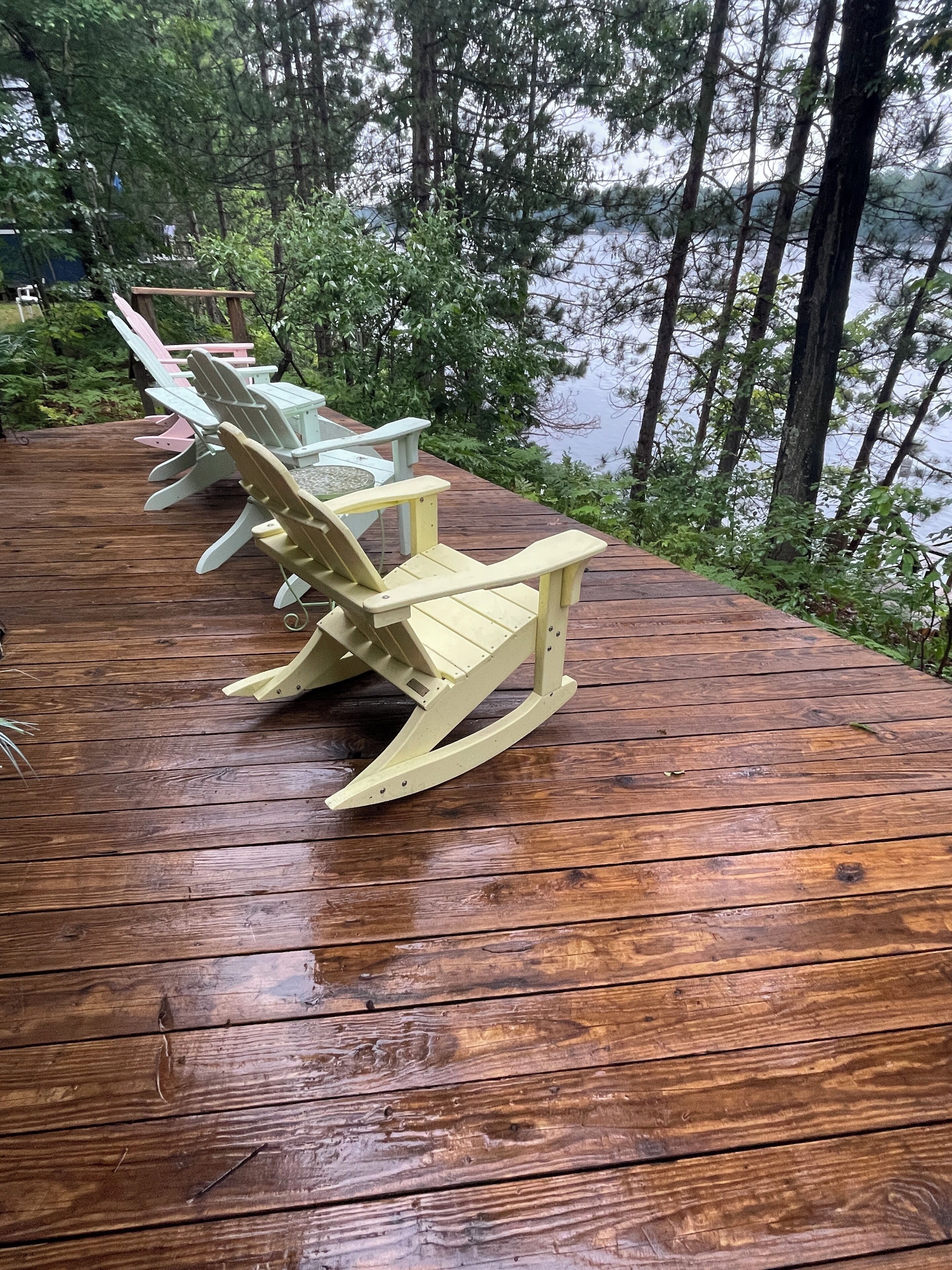 A wooden deck with Adirondack chairs and a table, surrounded by trees and a body of water.