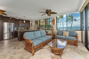 A modern kitchen with stainless steel appliances, a bar area, wicker furniture, and a large window with a view of palm trees and the ocean.