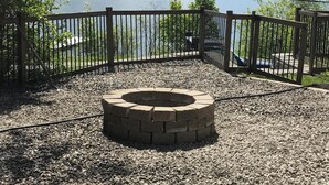 A gravel area with a circular fire pit, surrounded by a wooden fence, overlooking a lake and distant mountains.