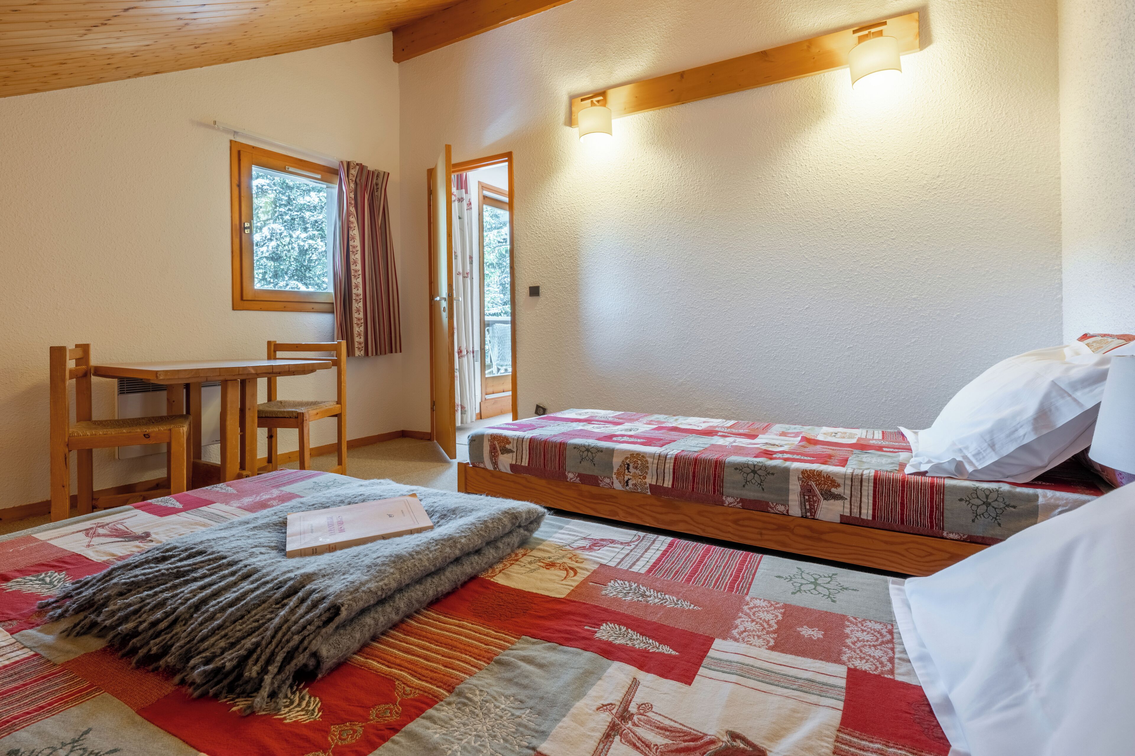A bedroom with a wooden bed, a small desk, and a chair. There is a window with curtains and a view of trees outside.