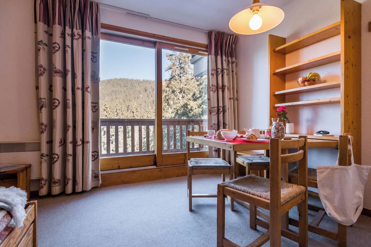 Een gezellige kamer met een houten eettafel, stoelen en uitzicht op een bos door grote ramen.
