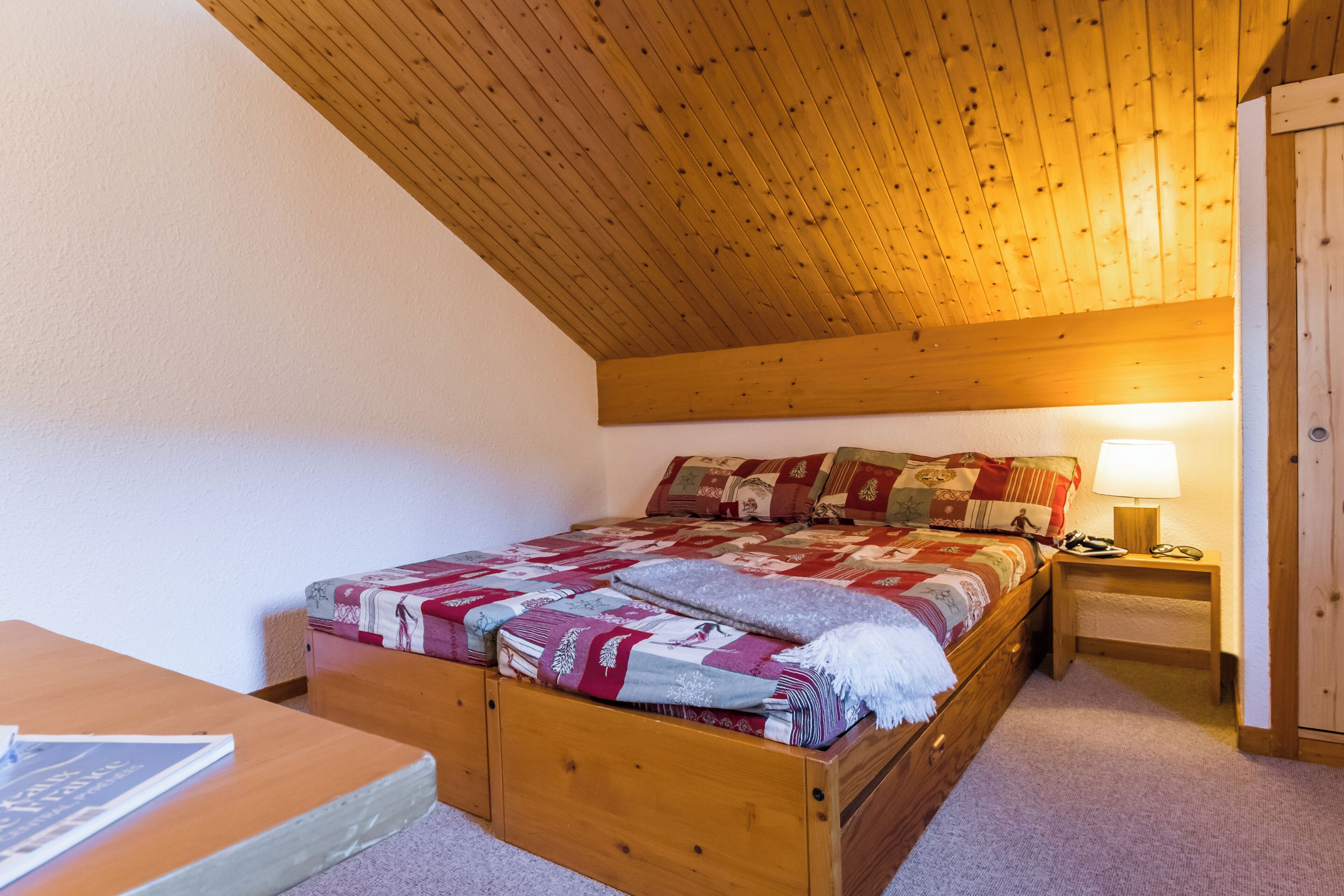 A bedroom with a wooden ceiling, a bed with a quilt, a bedside table with a lamp, and a nightstand.