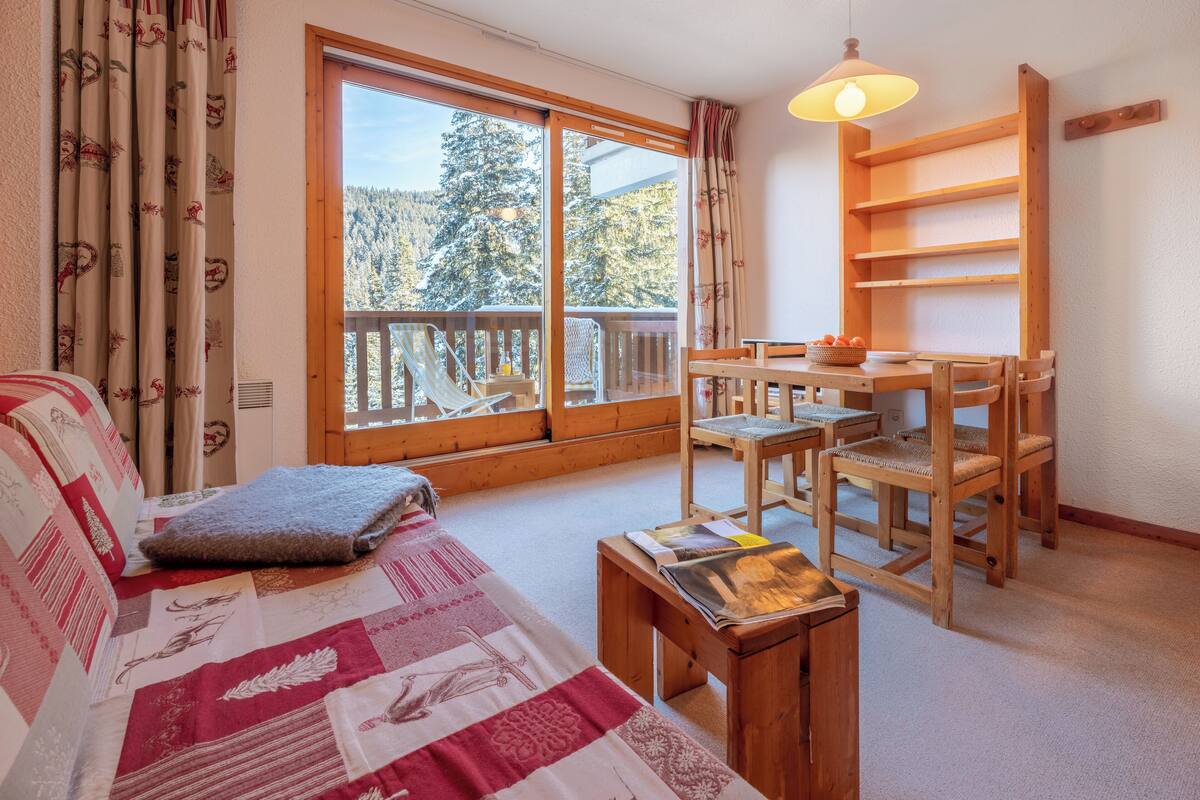 Een gezellige kamer met een bed, een houten tafel met stoelen, en een groot raam met uitzicht op bomen.