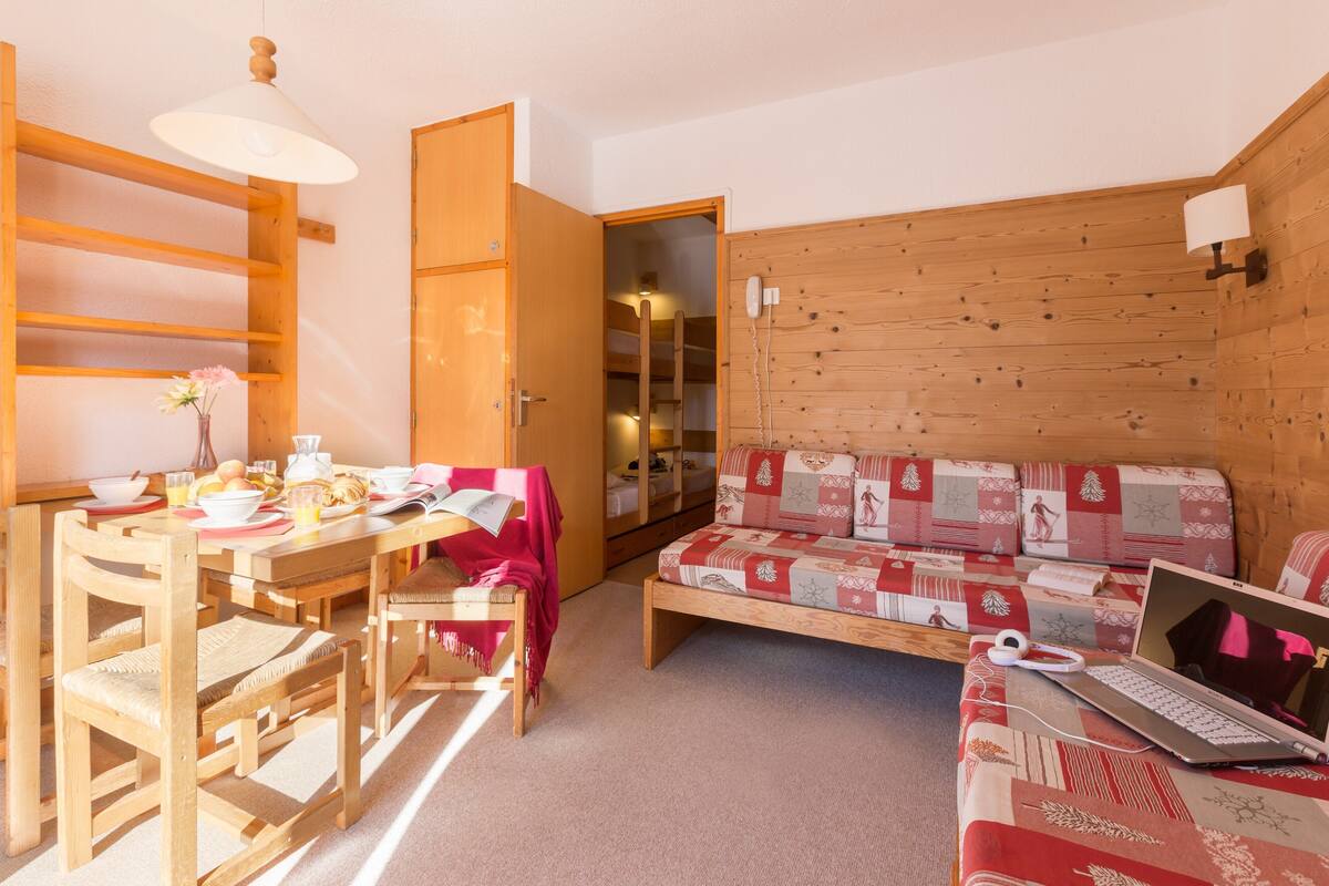 Een gezellige kamer met een houten eettafel gedekt voor het ontbijt, een bank met een gewatteerde hoes en een laptop op de bank.