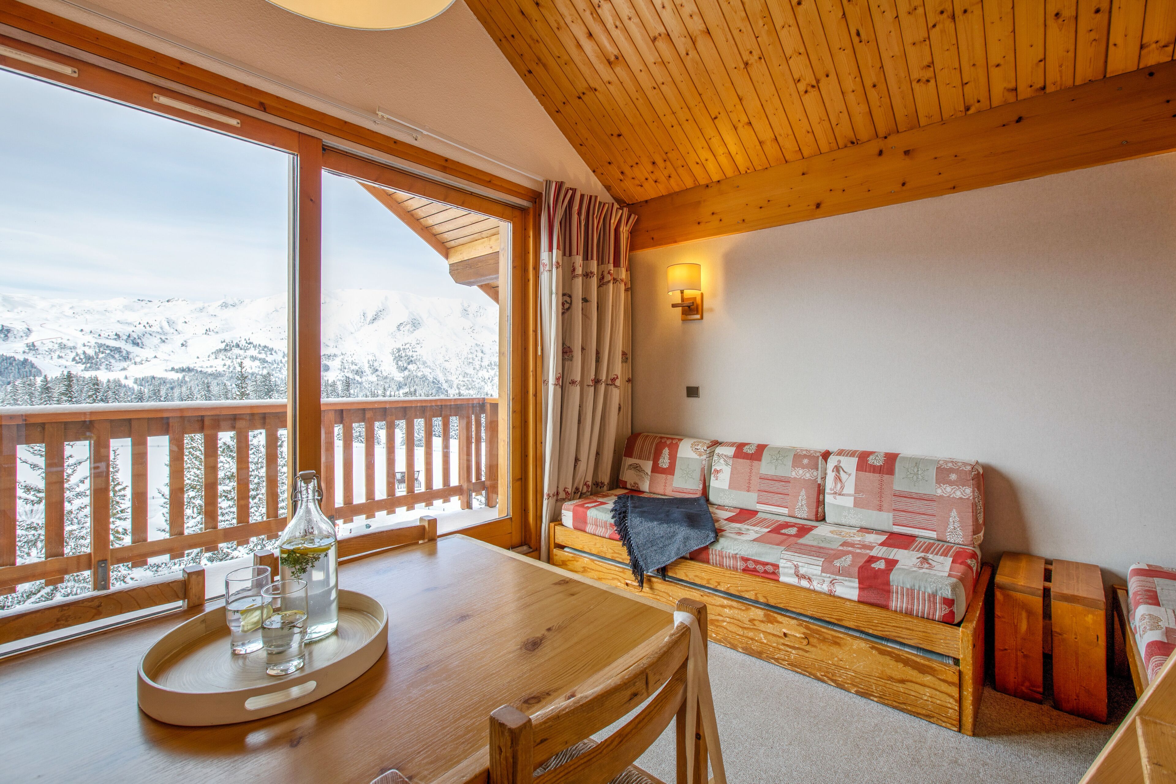 A wooden interior with a wooden table, a sofa, and a balcony with a view of snow-covered mountains.