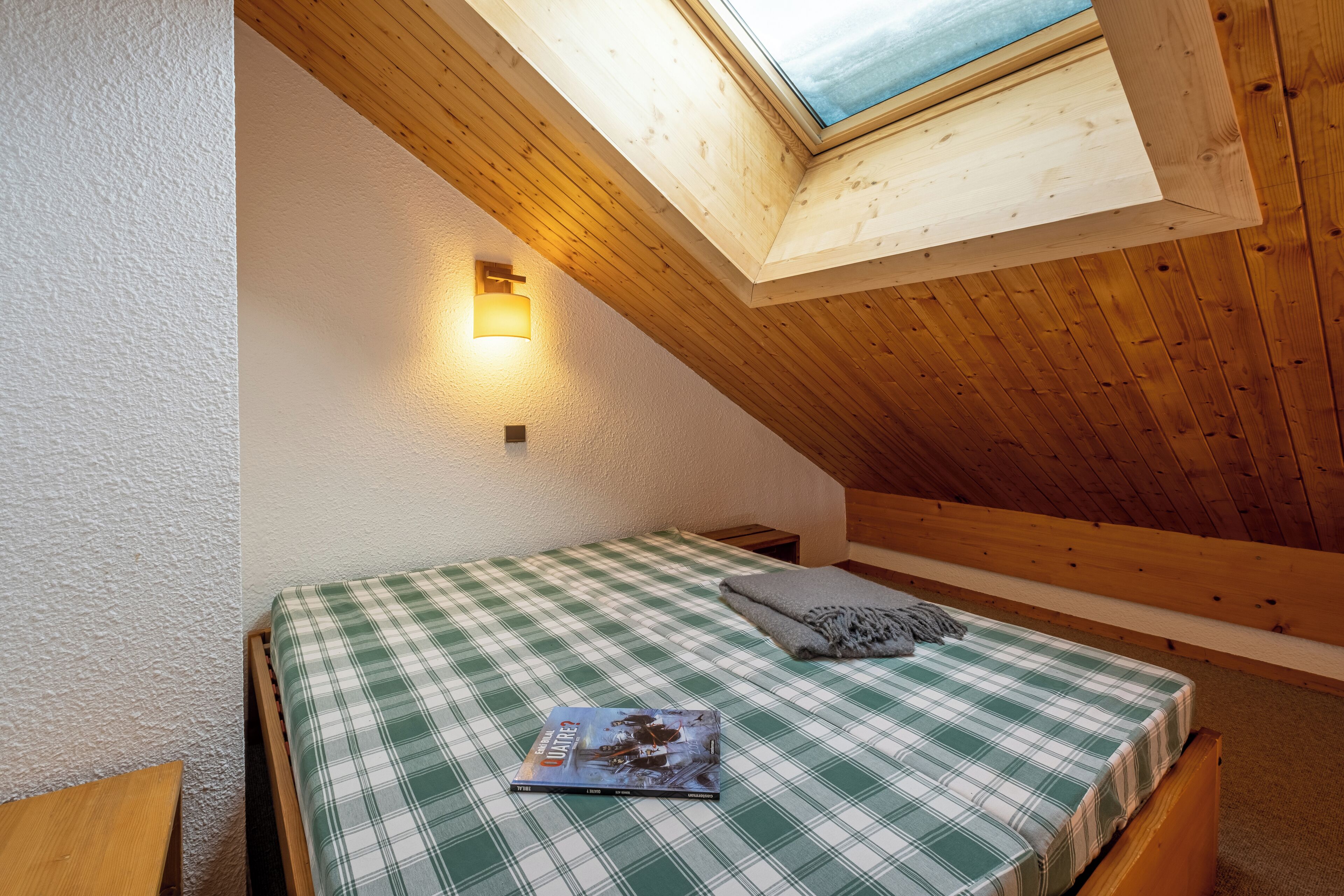 Una habitación pequeña y acogedora con techo inclinado y tragaluz. Se ve una cama individual con edredón de cuadros y una almohada, junto con una mesita de noche y una lámpara fijada a la pared.