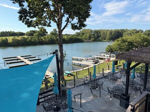 Eine Uferterrasse mit Tischen und Stühlen, ein Steg mit Booten und ein Gebäude mit einem Reetdach.