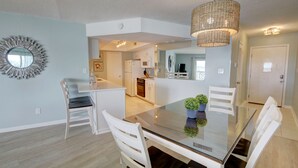 A modern dining area with a glass table, white chairs, a hanging light fixture, and a kitchen in the background.