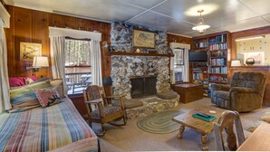 A cozy living room with a stone fireplace, wooden paneling, a sofa, a chair, a coffee table, and a bookshelf filled with books. - (80) Chatterton's Chalet at The Redwoods In Yosemite (Yosemite National Park)