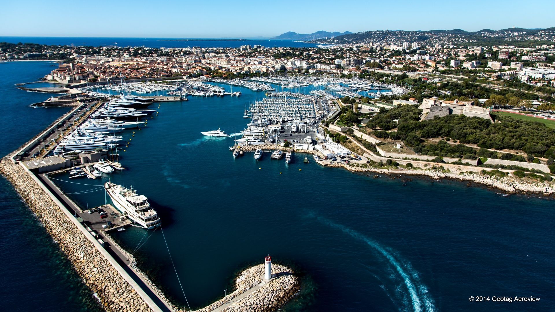 An aerial view of a marina with numerous boats docked, a lighthouse, and a coastal cityscape in the background.