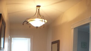Un couloir avec un plancher en bois, un canapé gris orné d’un coussin à motifs, un miroir fixé au mur et une porte donnant sur une chambre à coucher avec un lit.