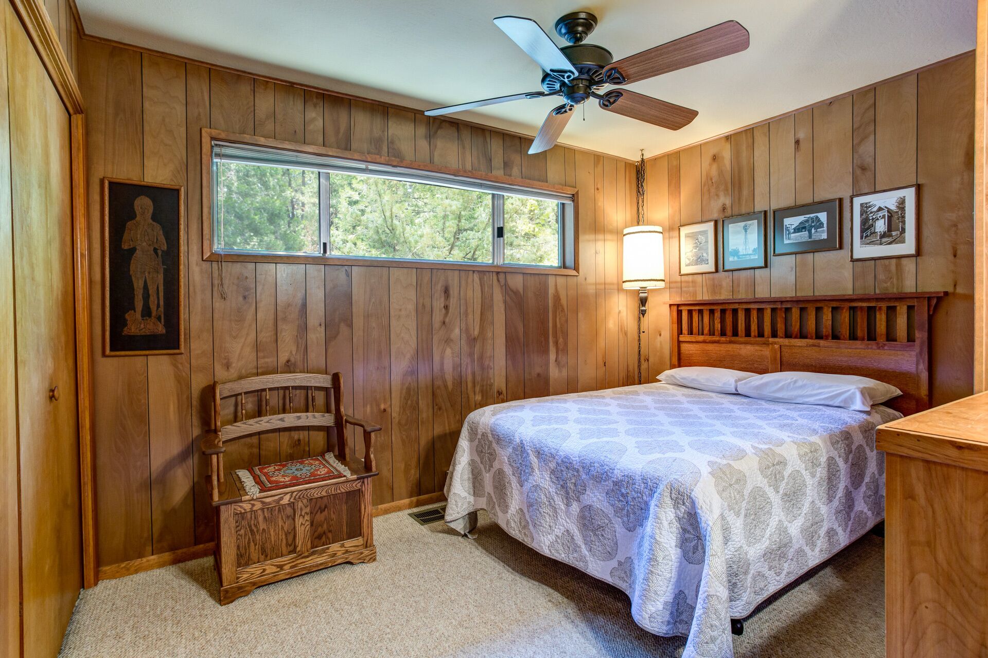 A bedroom with wooden walls, a bed with a patterned bedspread, a wooden bench, a ceiling fan, and a window with a view of trees.