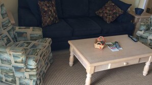 A living room with a dark blue sofa, patterned cushions, a wooden coffee table with remote controls, and a tiled floor. - Magnificent beach front condo with pool and restaurant/bar (Freeport)