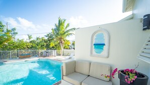 A poolside area with a beige cushioned bench, a black trash can, and a framed picture of a beach scene. - Fleur de Mer Villa Gracebay ( semi-detached) 2 bdrm -2 min drive from the beach! (Grace Bay)