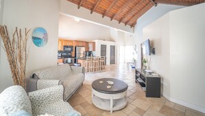 A spacious living area with a high ceiling, a sofa, a round coffee table, and a kitchen in the background. - Fleur de Mer Villa Gracebay ( semi-detached) 2 bdrm -2 min drive from the beach! (Grace Bay)