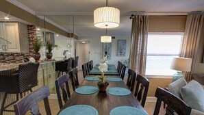 A dining area with a long wooden table, blue placemats, and chairs. There is a chandelier above the table and a kitchen area to the left.