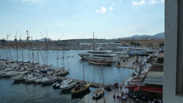 Un port de plaisance où sont amarrés différents types de bateaux, une jetée où des personnes se promènent, et des bâtiments en arrière-plan.