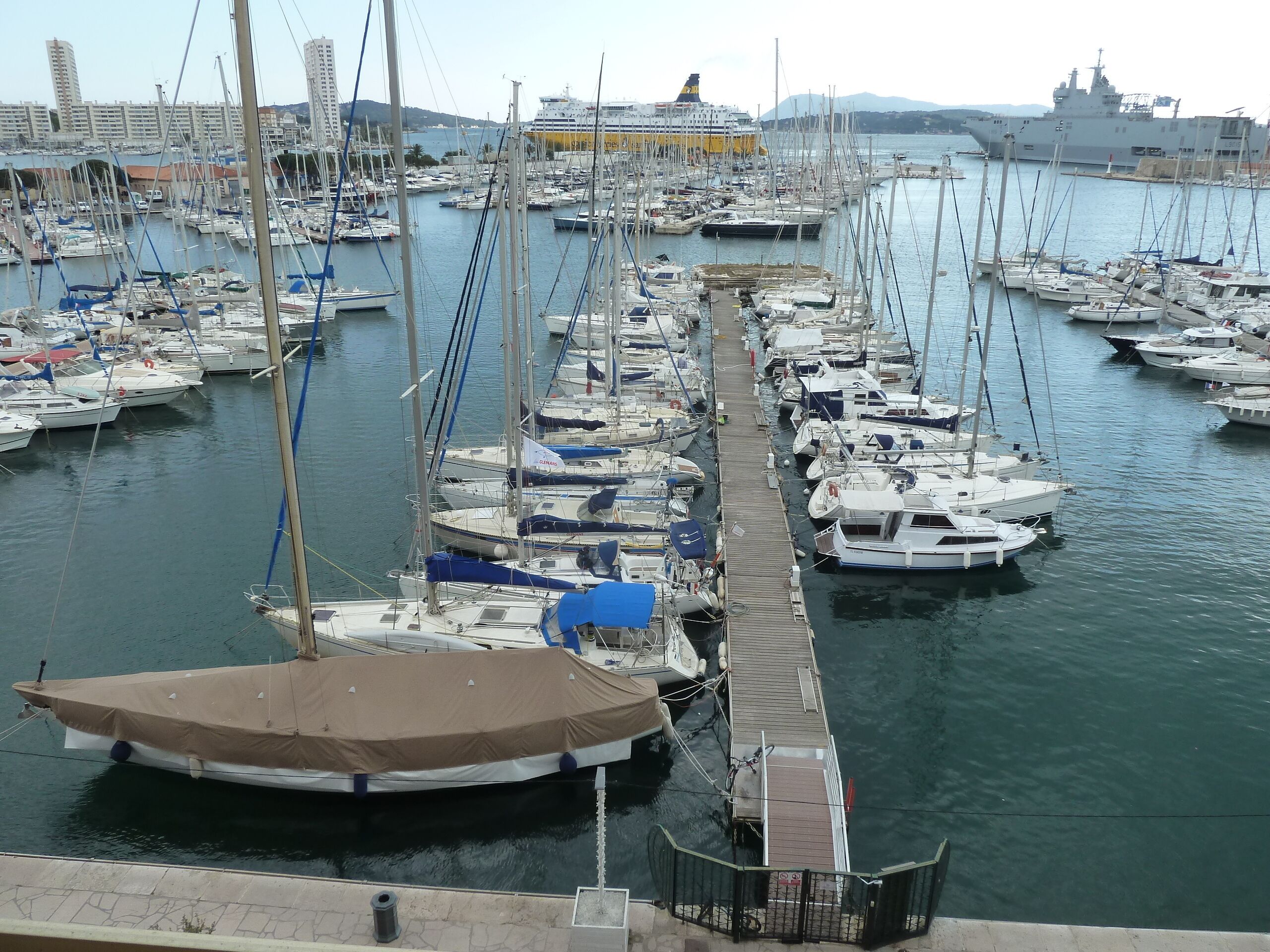 Un port de plaisance où sont amarrés de nombreux bateaux, un grand navire de croisière en arrière-plan et un paysage urbain avec des bâtiments et des collines.
