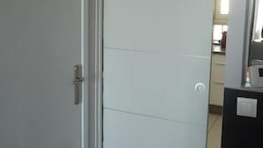 Une porte blanche fermée, dotée d’une poignée et de charnières apparentes, et laissant entrevoir une pièce dont on aperçoit un sol carrelé et une fenêtre.