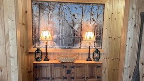 A wooden-paneled room with a central console table, two wall-mounted lamps, and a large framed painting of birds.
