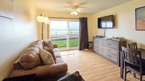 A living room with a sofa, a TV, a dining table with chairs, a ceiling fan, and a view of the beach through large windows.