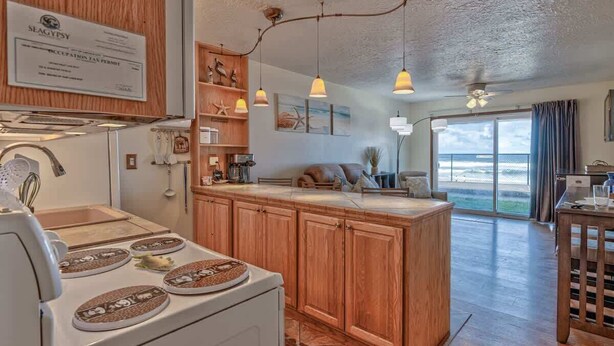 A kitchen with wooden cabinets, a white stove, and pendant lights. There is a certificate on the wall and a view of the ocean through large windows.