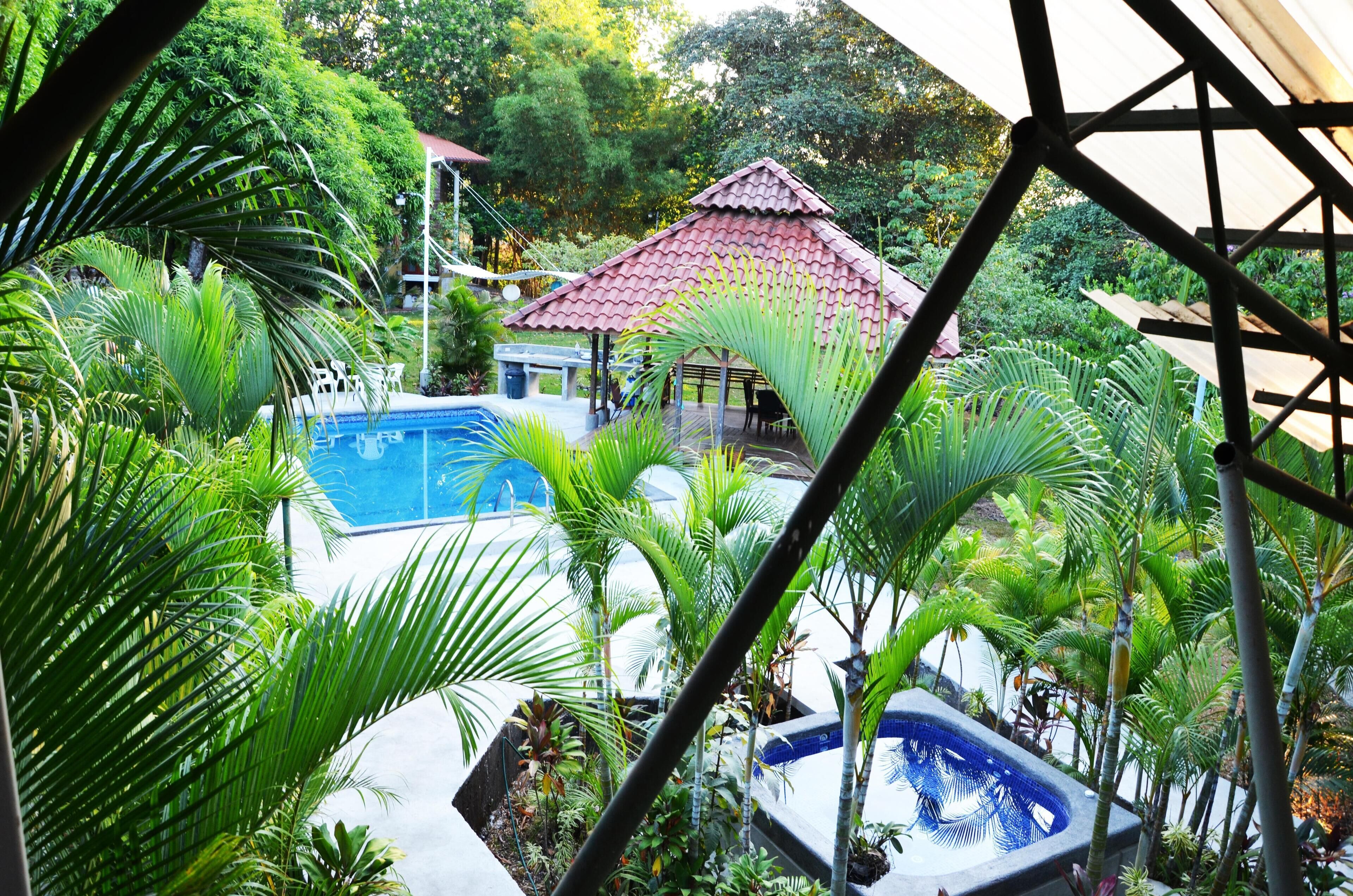 Un área de piscina rodeada de abundante vegetación, un pabellón y un pequeño edificio con techo rojo.