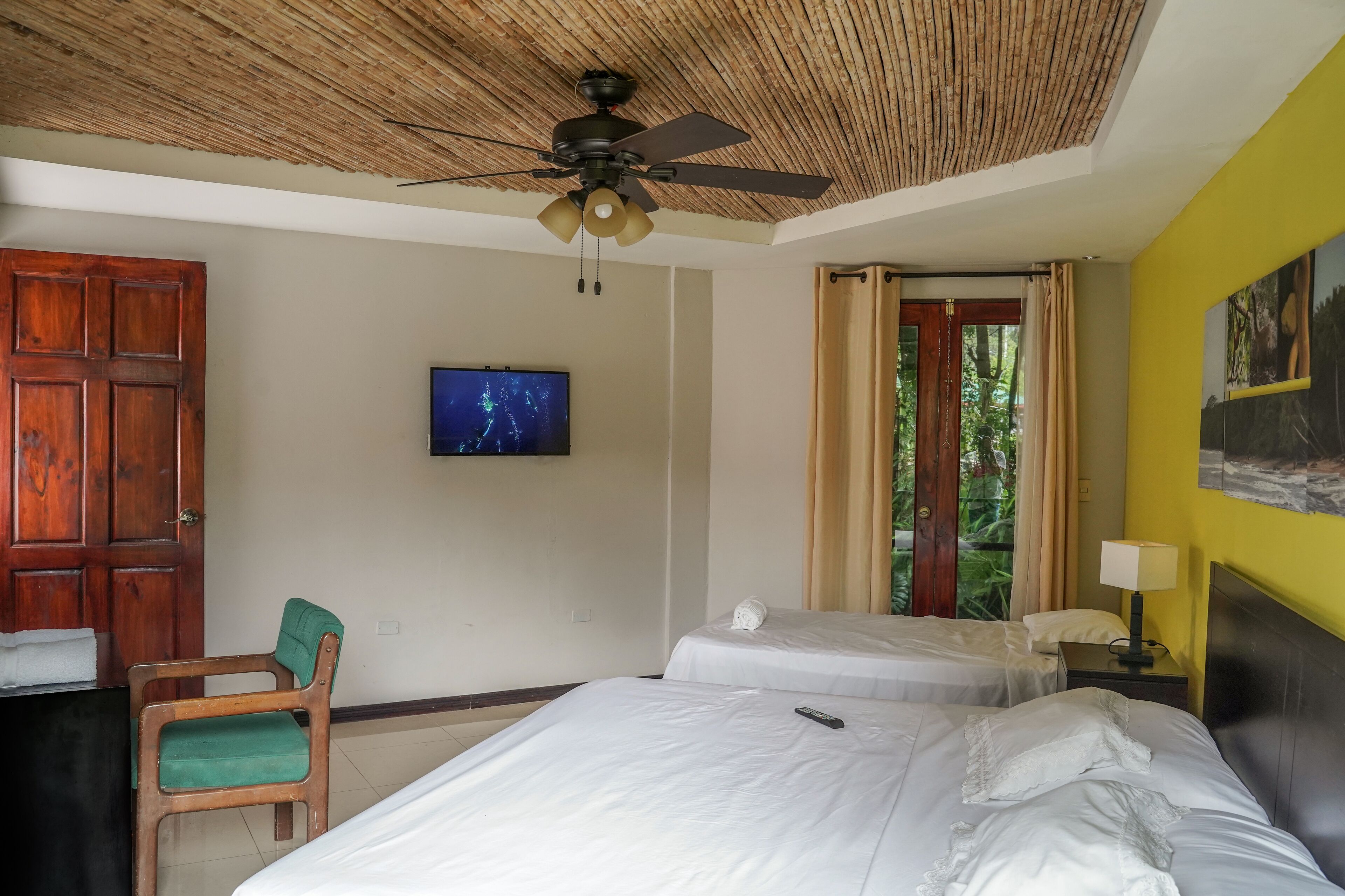 A hotel room with a ceiling fan, a TV, two beds, a chair, and a window with curtains.