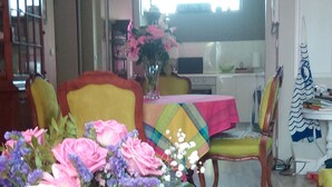 Un salon avec une table basse en bois, un vase rempli de fleurs, une table à manger recouverte d’une nappe colorée, et une cuisine en arrière-plan.