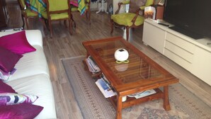 Un salon avec un canapé, des fauteuils, une table basse, un téléviseur et, au fond, un coin cuisine.